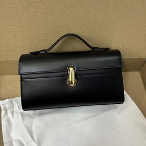 May include: A black handbag with a top handle and a gold-colored clasp. The rectangular bag has a structured design and a flap closure. The bag is sitting on a white cloth inside a cardboard box.
