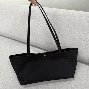 May include: A black rectangular tote bag featuring a smooth fabric construction, two long thin shoulder straps, and a single silver-toned metal snap button closure centred at the top opening of the bag for secure storage of personal items.