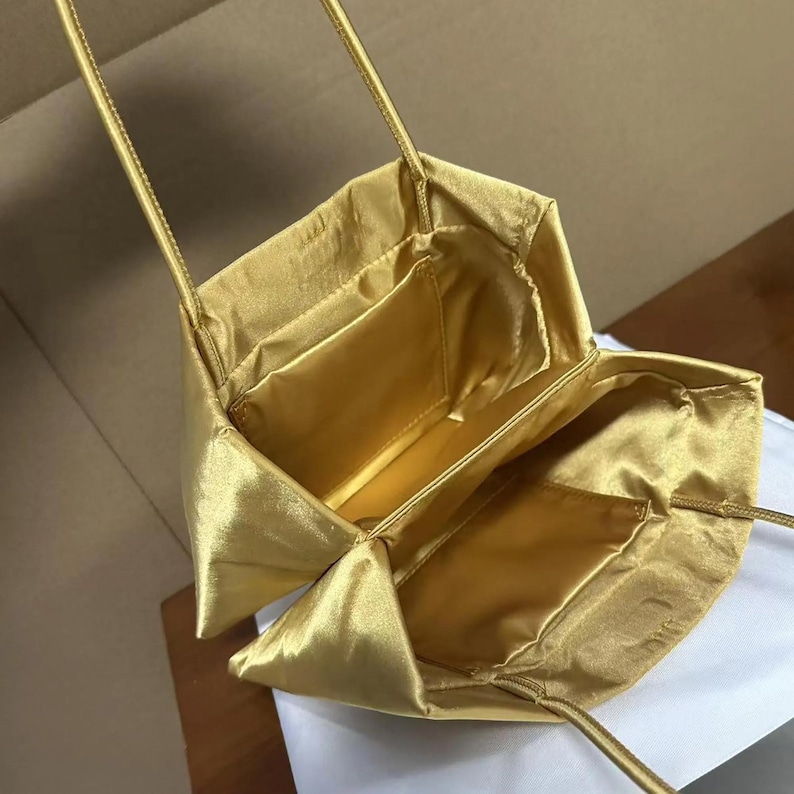 May include: A gold satin handbag with a structured, open-top design and thin shoulder straps. The interior features a smooth, matching gold lining with a single flat pocket on the inner wall, showcasing a lustrous, reflective fabric finish.