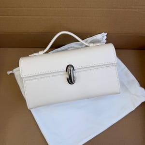 May include: Cream-colored rectangular handbag with a top handle and a silver-colored clasp. The bag is sitting on a white cloth inside a cardboard box. The handbag has a structured design and a minimalist aesthetic.