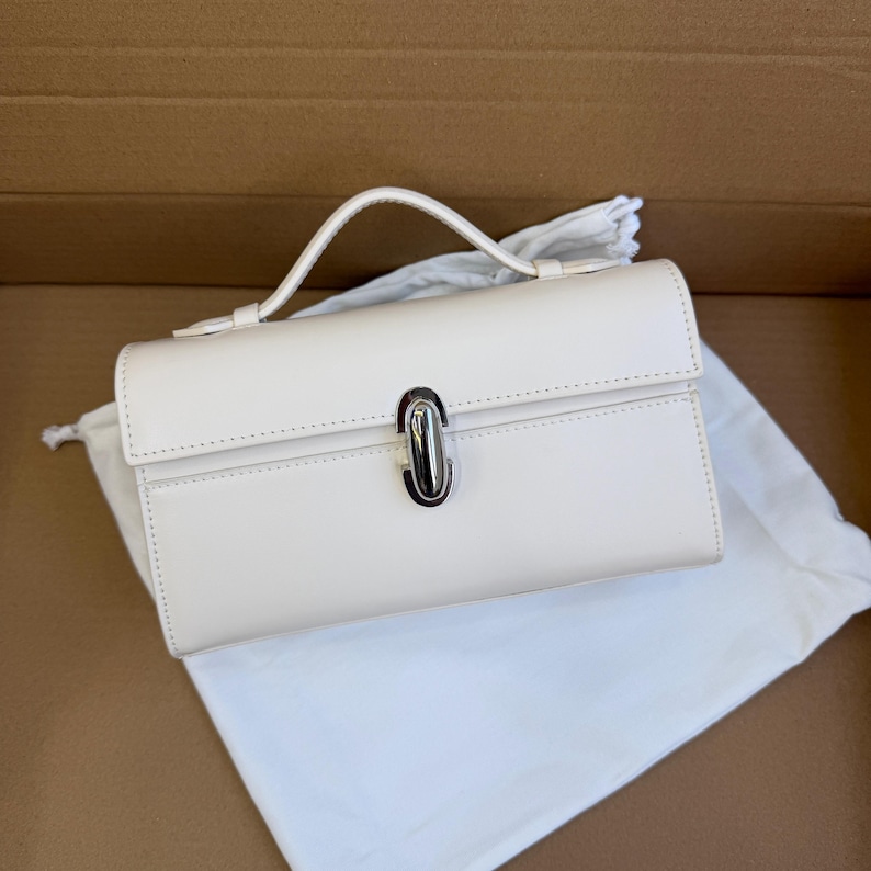 May include: Cream-colored rectangular handbag with a top handle and a silver-colored clasp. The bag is sitting on a white cloth inside a cardboard box. The handbag has a structured design and a minimalist aesthetic.