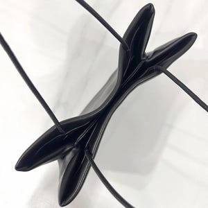 以下が含まれることがあります： 滑らかでマットな質感の黒いショルダーバッグ。構造的で折り畳まれたデザインが特徴です。上部の縁には2本の細いコード状のストラップが付いており、上から見るとV字型の開口部が形成され、バッグの暗い内部が見えるようになっています。