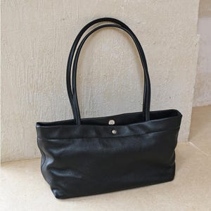 Peut inclure: Un sac cabas rectangulaire noir à la texture lisse, doté de deux longues anses d'épaule et d'une fermeture à bouton-pression magnétique argentée au centre supérieur. Le sac présente une base structurée et un design épuré sans poches ni logos.