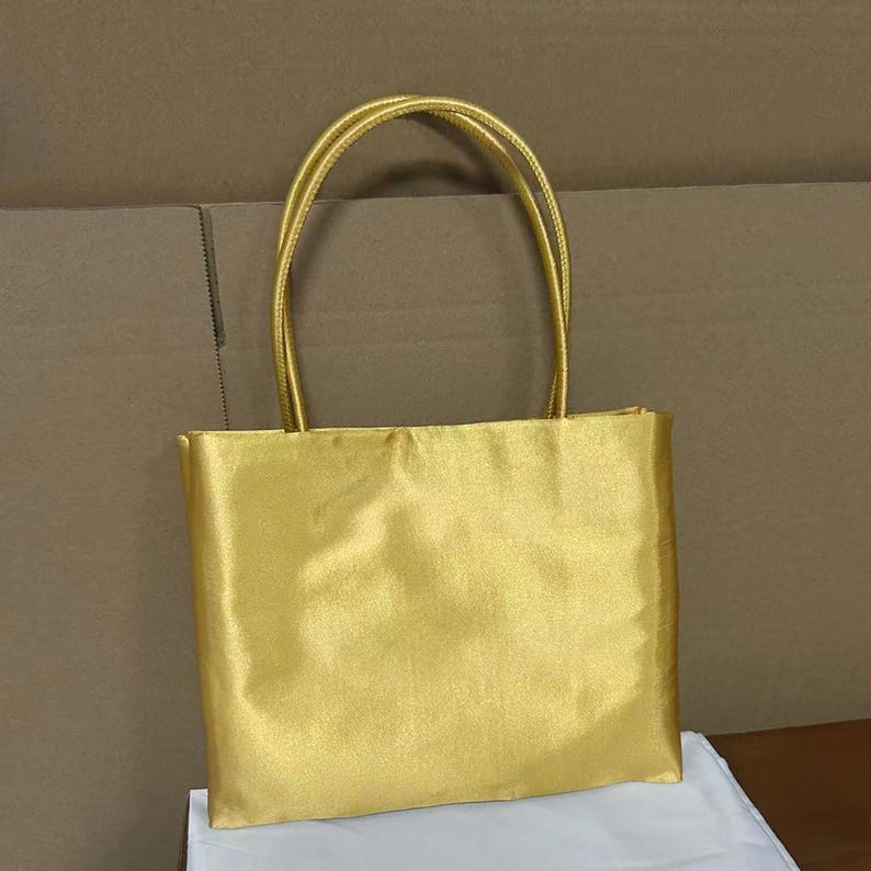 May include: A rectangular tote bag crafted from a shimmering gold satin fabric. The bag features two matching gold fabric handles attached to the top edge, providing a structured yet soft appearance with a smooth, reflective surface texture.
