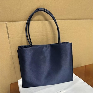 May include: A rectangular navy blue handbag with a smooth, satin-like finish. The bag features two thin, matching navy blue shoulder straps attached to the top edge. The design is simple and structured, with a clean front panel and no visible hardware.