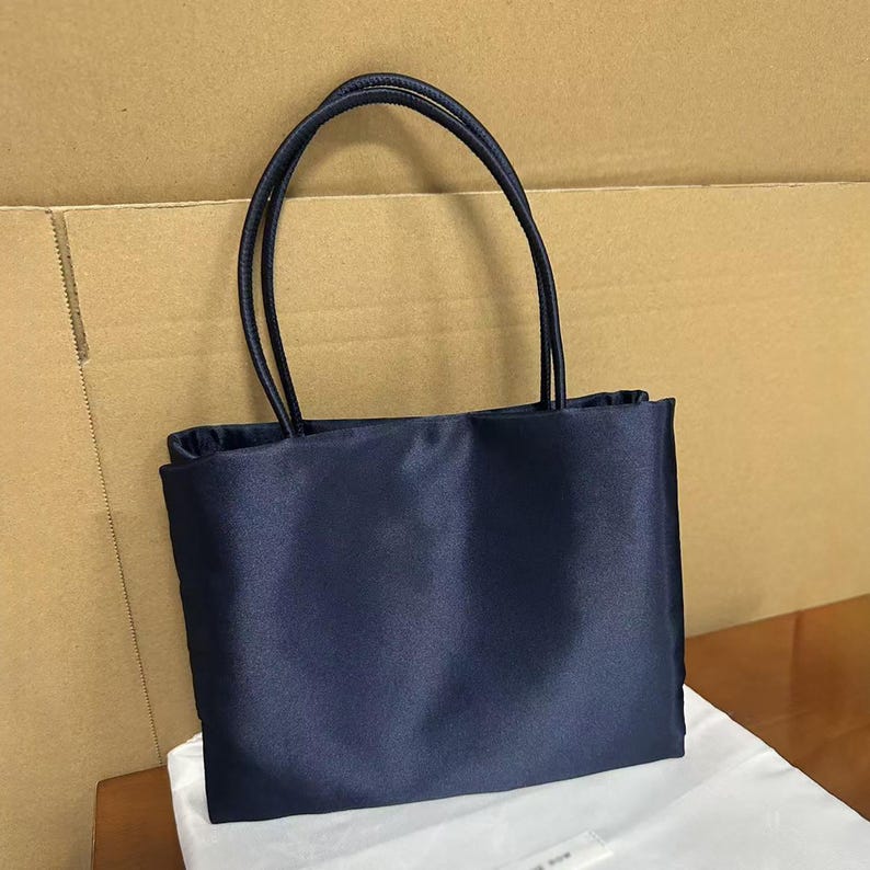 May include: A rectangular navy blue handbag with a smooth, satin-like finish. The bag features two thin, matching navy blue shoulder straps attached to the top edge. The design is simple and structured, with a clean front panel and no visible hardware.