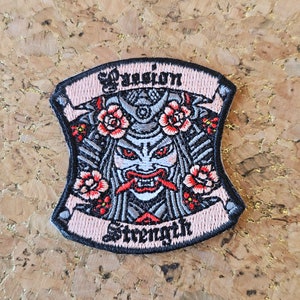 May include: Embroidered patch with a pink background and black outline. The patch features a stylized image of a Japanese demon with red eyes and fangs, surrounded by pink flowers. The text "Passion" and "Strength" is embroidered in black on the banner.