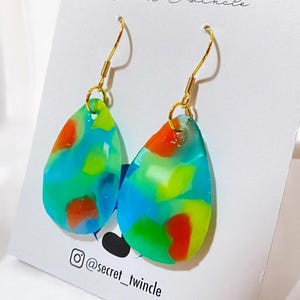 May include: A pair of teardrop-shaped earrings with a colorful abstract design. The earrings feature a mix of blue, green, and orange hues, with gold-colored ear wires. The earrings are displayed on a white card with the text "Secret Twincle".
