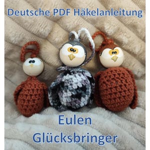 Op de afbeelding: Drie handgemaakte uilenornamenten. De uilen hebben beschilderde houten hoofden met gele ogen en snavels. De lichamen zijn gehaakt in bruin-, grijs- en wittinten. De tekst "Deutsche PDF Häkelanleitung" en "Eulen Glücksbringer" is zichtbaar.