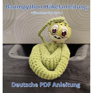 Op de afbeelding: Een gehaakt groen slangenknuffel met een beschilderde houten kop. De kop heeft grote ogen en een lachende uitdrukking. De tekst "Baumpython Häkelanleitung" en "Deutsche PDF Anleitung" zijn zichtbaar.