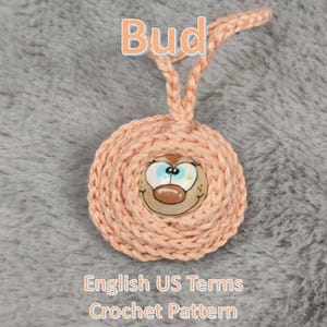 May include: A peach-colored crocheted ornament with a cartoon face in the center. The ornament has a loop at the top for hanging. The word "Bud" is written at the top of the image.