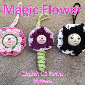Könnte beinhalten: Drei handgefertigte Häkelblumen-Ornamente in Rosa, Lila und Schwarz. Jede Blume hat einen Knopf mit lächelndem Gesicht in der Mitte und eine Schlaufe zum Aufhängen. Der Text "Magic Flower" und "English US Terms Pattern" sind sichtbar.
