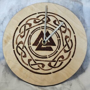 Peut inclure: Une horloge ronde en bois clair. Le cadran présente un motif gravé brun foncé d'un triangle dans un cercle, entouré d'un motif de nœuds complexe. Les aiguilles argentées indiquent l'heure.