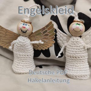 May include: Two handmade angel ornaments. Each angel has a wooden bead head with painted facial features, a gold halo, and white crochet body. One angel has gold-colored wings, the other has white wings. The text "Engelskleid" and "Deutsche PDF Häkelanleitung" are visible.