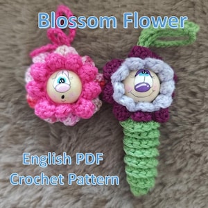 Könnte beinhalten: Zwei gehäkelte Blumenornamente mit bemalten Gesichtern. Die rosa Blume hat eine rosa und weiße gehäkelte Blume mit einem weißen Gesicht und blauen Augen. Die lila Blume hat eine lila und graue gehäkelte Blume mit einem weißen Gesicht und lila Augen. Die Blumen sind an grünen gehäkelten Stielen befestigt. Der Bildtext lautet "Blossom Flower English PDF Crochet Pattern".