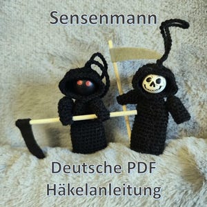 May include: Two handmade crochet figures of the Grim Reaper. One figure has red eyes and a scythe, the other has a skull face and a flag. The figures are black with hoods. Text on the image reads "Sensenmann" and "Deutsche PDF Häkelanleitung".