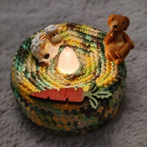 Op de afbeelding: Een handgemaakte gehaakte kandelaar met een schaap- en berenfiguur. De houder is in tinten geel, groen en bruin. Een houten wortel is aan de voorkant bevestigd. Er zit een LED-kaars in.