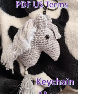 Peut inclure: Un porte-clés cheval gris crocheté avec un pompon argenté et un œil noir. Le porte-clés est attaché à un anneau argenté. Le texte "Crochet Pattern PDF US Terms" et "Keychain Horse" sont visibles.
