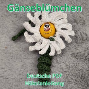 May include: A handmade crochet daisy with a yellow center featuring a smiling face. The white petals surround the yellow center. The stem is green. Text in the image reads "Gänseblümchen" and "Deutsche PDF Häkelanleitung."