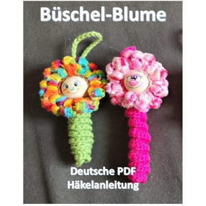 May include: Two handmade crochet flower ornaments. One has a colorful rainbow flower with a green stem and loop. The other has a pink flower with a pink stem. The flowers have a smiling face in the center. Text at the top says "Büschel-Blume" and at the bottom "Deutsche PDF Häkelanleitung".