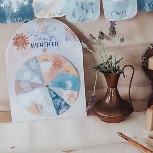 May include: A wooden weather wheel with a colorful illustration of different weather conditions, including sun, rain, snow, and wind. The wheel is surrounded by a white card with the text "Today's Weather". There is a rainbow painted on a white surface with paint pots and a paintbrush.