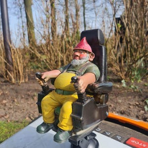 May include: A garden gnome figurine in a black chair, wearing a red hat and yellow trousers, operating controls. The gnome has a beard and is wearing a green shirt. The figurine is set outdoors, possibly in a garden or yard.