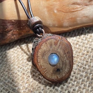 Op de afbeelding: Een handgemaakte hangerketting met een ronde houten hanger met een natuurlijke boomschorsrand. De hanger heeft een centrale, lichtblauwe, maansteenachtige edelsteen. De ketting heeft een bruin leren koord en een houten sluiting. De hanger is ongeveer 3,8 cm in diameter.
