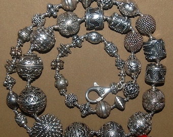May include: A silver necklace with a variety of beads, including round, cylindrical, and textured beads. The necklace has a silver clasp and a single red bead.