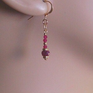 May include: A gold-colored earring with a hook, featuring a dangling design. The earring showcases a series of small, round, ruby-colored beads, interspersed with gold-colored beads, culminating in a larger, faceted ruby-colored bead at the bottom.