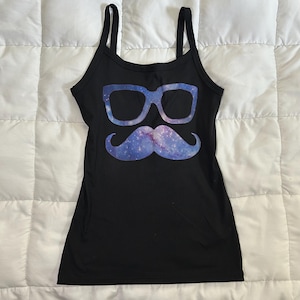 May include: A black tank top featuring a graphic print of eyeglasses and a mustache. Both the glasses and the mustache are filled with a blue and purple galaxy pattern that includes small white stars and nebulae against the solid black fabric.