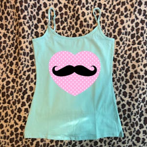 May include: A light mint green camisole tank top featuring thin adjustable spaghetti straps and a scoop neckline. Centered on the front is a large pink heart graphic with a white polka dot pattern and a solid black handlebar mustache icon.