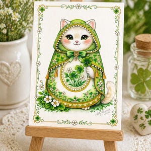 May include: A whimsical illustration of a white cat dressed in a green hooded cloak, holding a four-leaf clover. The cat is adorned with shamrocks and white flowers, set against a white background with a decorative border. The artwork is displayed on a small wooden easel.