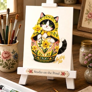 May include: A whimsical watercolor painting of a cat dressed as a Matryoshka doll, holding daffodils. The artwork is displayed on a small white easel with the text "Studio on the Pond". The cat is black and white with a yellow head covering.