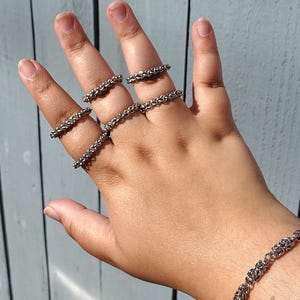 May include: A hand wearing multiple silver chain rings and a silver chain bracelet. The rings are stacked on each finger.