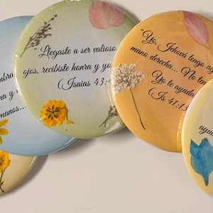 May include: A collection of five round, decorative magnets with floral accents and Spanish text. Each magnet features a different pastel color, dried flowers, and a handwritten quote from the Bible. The magnets are a unique, inspirational home decor item.