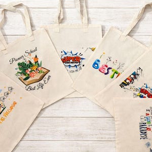 May include: A collection of off-white canvas tote bags with various colorful graphic designs. One bag features an open book with flowers, another says "SUPER", and others have text like "BEST" and "I AM A PARADISE".