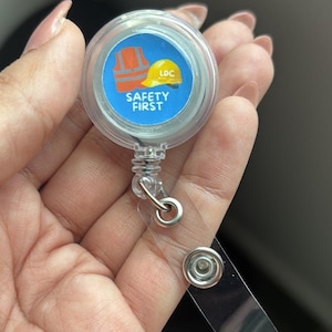 May include: A clear, retractable badge reel with a blue and white graphic that reads "SAFETY FIRST." The graphic includes a yellow hard hat and an orange safety vest. The reel has a silver metal clip and a clear plastic strap.