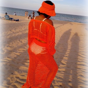 May include: A woman wearing a bright orange crochet maxi dress with a matching crochet bucket hat. The dress has a fringe hem and is worn over a white crochet bikini top. The woman is standing on a sandy beach with the ocean in the background.