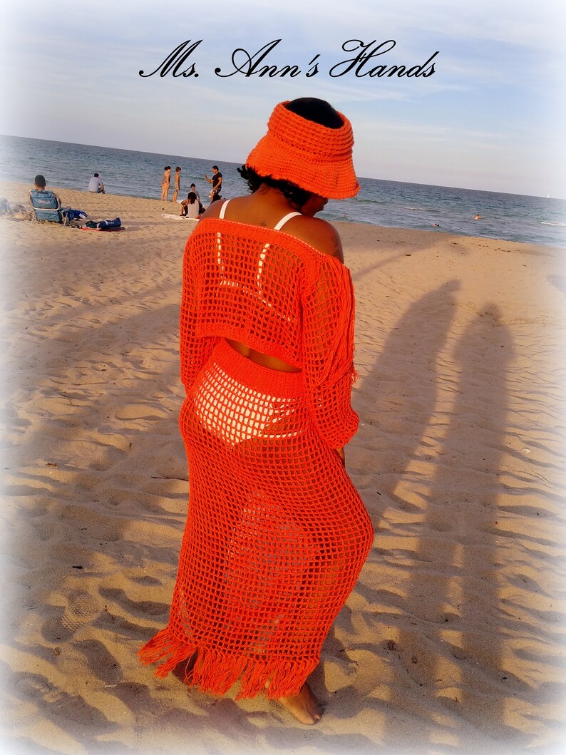 May include: A woman wearing a bright orange crochet maxi dress with a matching crochet bucket hat. The dress has a fringe hem and is worn over a white crochet bikini top. The woman is standing on a sandy beach with the ocean in the background.