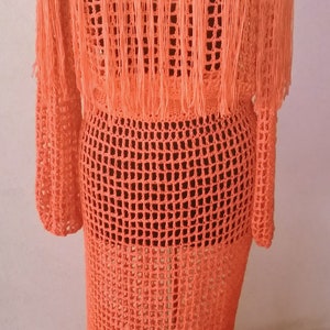 May include: A long, orange crochet dress with fringe detailing. The dress is made of a lightweight, open-weave fabric. "Ms. Ann's Hands" is written on the hanger.