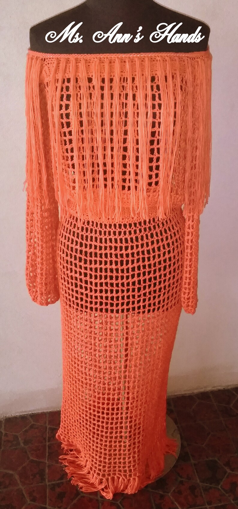 May include: A long, orange crochet dress with fringe detailing. The dress is made of a lightweight, open-weave fabric. "Ms. Ann's Hands" is written on the hanger.