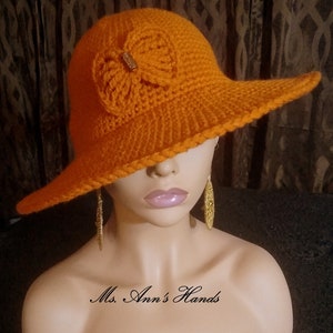 May include: A bright orange crochet sun hat with a large brim and a crocheted bow detail. The hat is on a mannequin's head. The text "Ms. Ann's Hands" is visible at the bottom of the image.