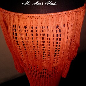 May include: Orange crochet skirt with fringe detail. The skirt is made with a loose, open weave and has a long, flowing silhouette.