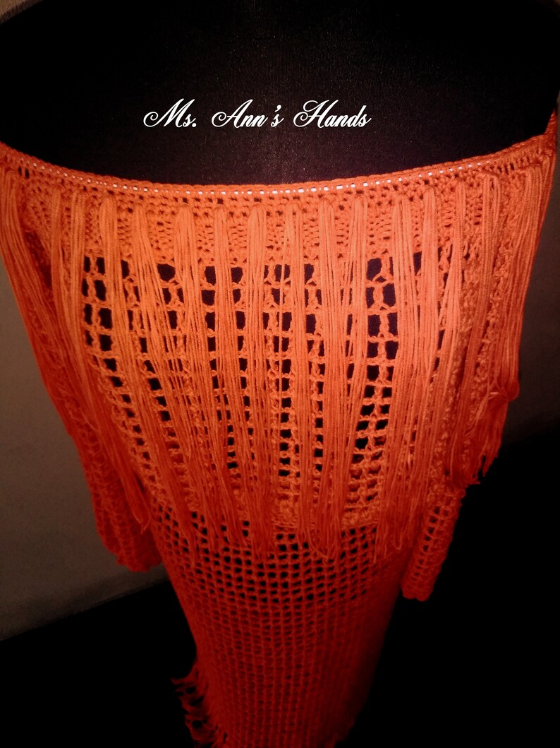 May include: Orange crochet skirt with fringe detail. The skirt is made with a loose, open weave and has a long, flowing silhouette.