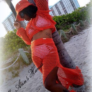 May include: Orange crochet beach cover-up with a matching hat. The cover-up has a long skirt with a high slit and a cropped top. The hat is a wide-brimmed sun hat with a crocheted design. The model is standing on a beach with a palm tree in the background. The text "Ms. Ann's Hands" is visible in the lower left corner of the image.