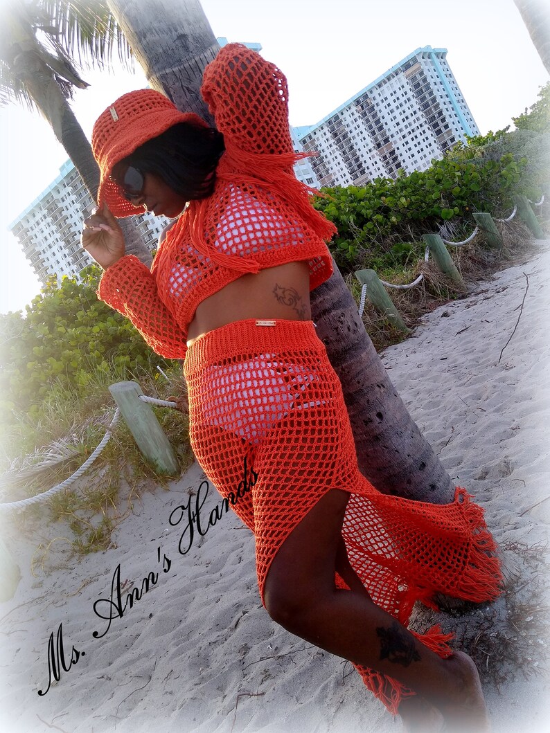 May include: Orange crochet beach cover-up with a matching hat. The cover-up has a long skirt with a high slit and a cropped top. The hat is a wide-brimmed sun hat with a crocheted design. The model is standing on a beach with a palm tree in the background. The text "Ms. Ann's Hands" is visible in the lower left corner of the image.