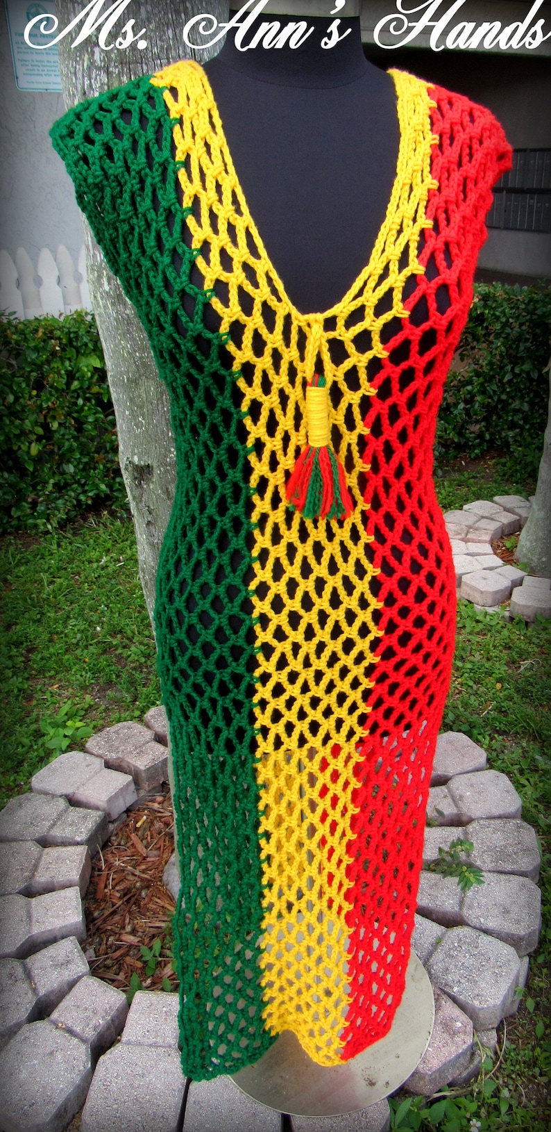 Peut inclure: Une robe en crochet de couleur verte, jaune et rouge. La robe a un col en V et un pompon au centre.