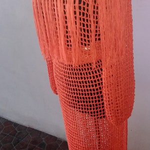 May include: Orange crochet dress with fringe detail. The dress is made of a lightweight, breathable fabric and has a loose, flowy fit.  "Ms. Ann's Hands" is written on the top of the dress.