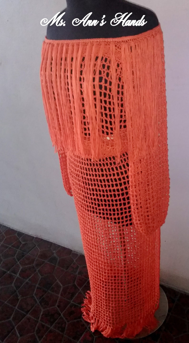 May include: Orange crochet dress with fringe detail. The dress is made of a lightweight, breathable fabric and has a loose, flowy fit.  "Ms. Ann's Hands" is written on the top of the dress.