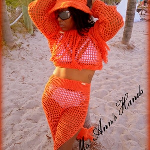 May include: Orange crochet top and trousers set with fringe detailing. The top is a cropped style and the trousers are wide-leg. The model is wearing a white crocheted hat and sunglasses. The text "Mrs. Ann's Hands" is visible in the image.